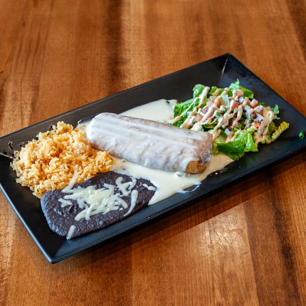 A burrito covered in white sauce, with rice, refried beans, and salad on a black plate.