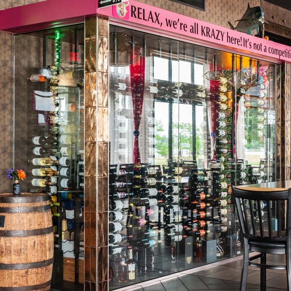 A glass-enclosed wine cellar with a pink sign reading "RELAX, we're all KRAZY here! It's not a competition!"
