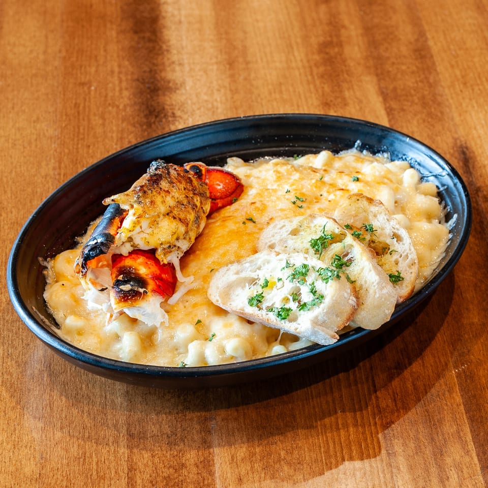 Lobster mac and cheese with toasted bread slices and parsley in a black bowl on a wooden table.
