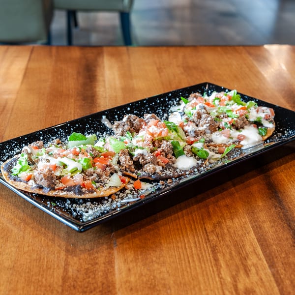 Three tostadas with ground beef, lettuce, tomatoes, and sour cream on a black rectangular plate.