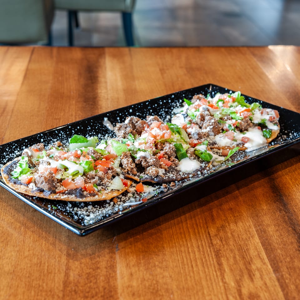 Three tostadas with ground beef, lettuce, tomatoes, and sour cream on a black rectangular plate.