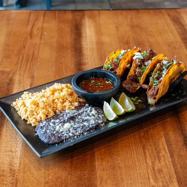 Four birria tacos with rice, refried beans, limes, and a dipping sauce on a black plate.