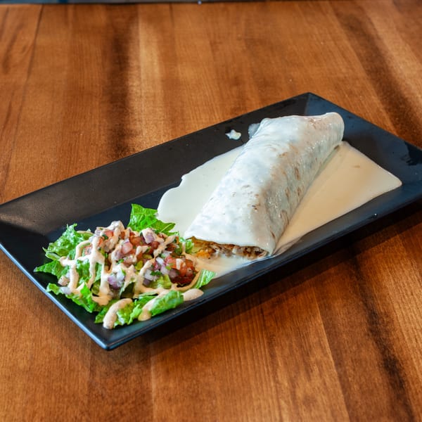 A large burrito covered in white sauce, served with a side salad on a black rectangular plate.