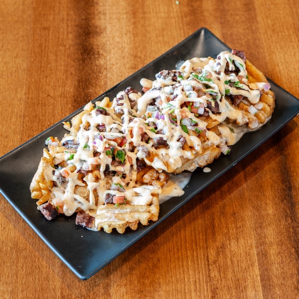 Waffle fries topped with steak, pico de gallo, and a creamy white sauce on a black plate.