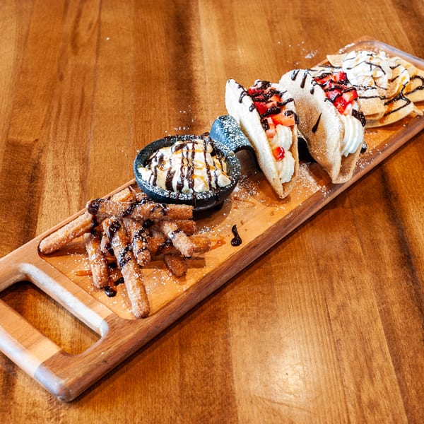 Dessert platter with churros, ice cream, dessert tacos, and mini pancakes on a wooden board.