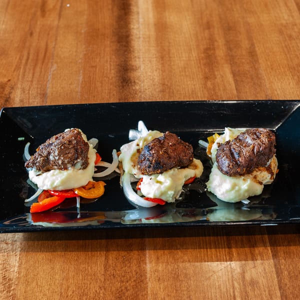 Three portions of meat and mashed potatoes with peppers and onions on a black rectangular plate.