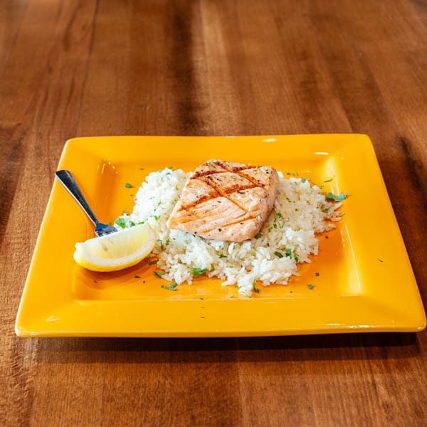 Grilled salmon on a bed of white rice with parsley, a lemon wedge, and a fork on a yellow plate.