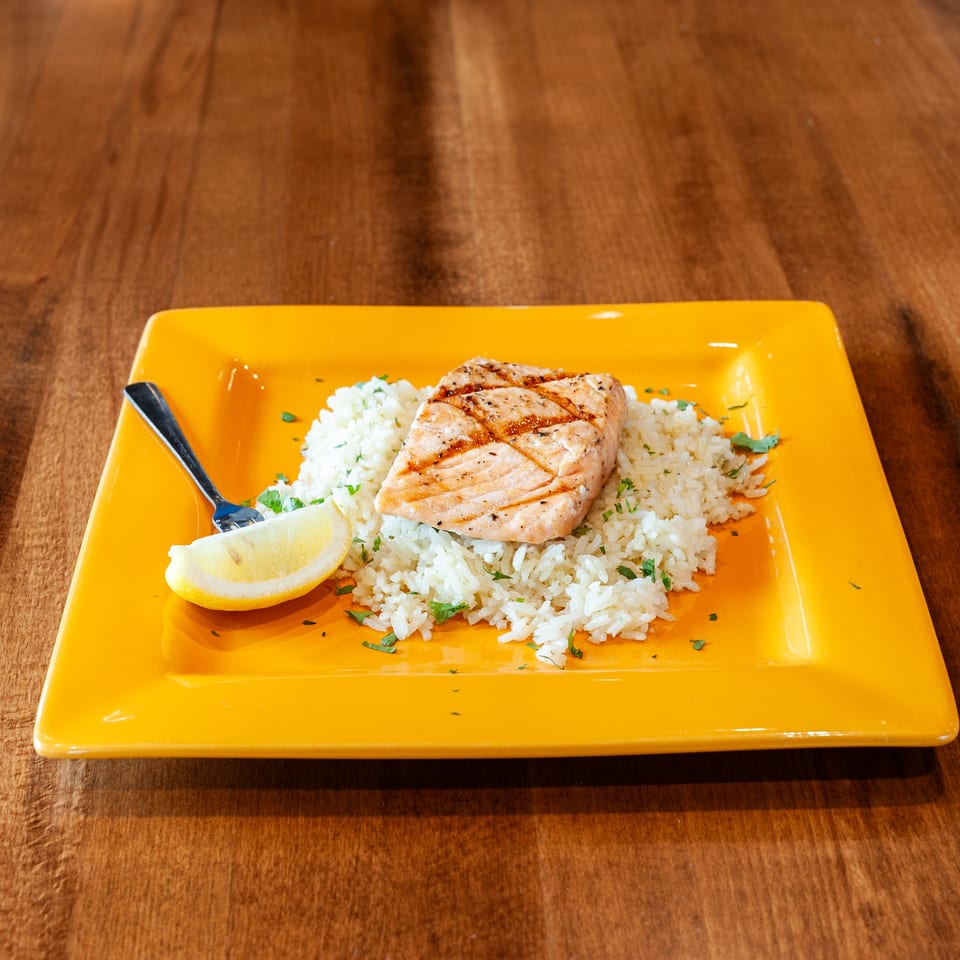 Grilled salmon on a bed of white rice with parsley, a lemon wedge, and a fork on a yellow plate.